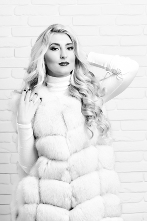 young fashionable sexy pretty woman with beautiful long curly blonde hair in waist coat of white fur and fashion makeup on brick wall studio backgroundの写真素材