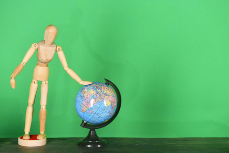Human shaped toy made of wood stands by little globe model. Geography lesson equipment and wooden statuette on green background, copy space. School items and whole world trip conceptの写真素材