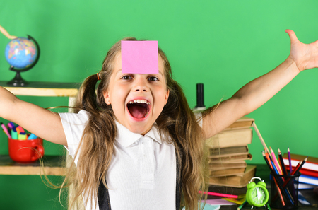 Back to school and childhood concept. Kid and school supplies on green wall background. Schoolgirl with cheerful face with pink sticker on forehead. Girl with ponytails stands by school itemsの写真素材
