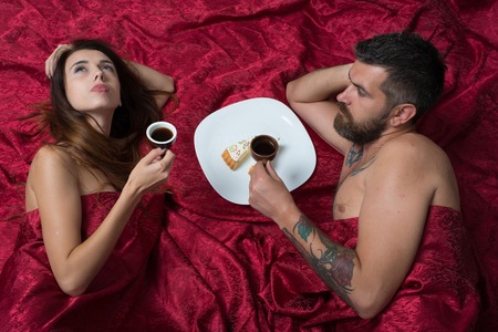 Man with beard having romantic breakfast with pretty lady in bed, top view. Love and sex concept. Man and woman with half covered bodies. Couple in love lies on burgundy sheets.の写真素材