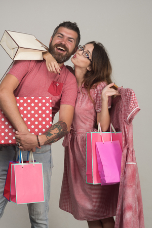Bearded man dressed in pink smiles. Couple holding shopping bags and dress, hugging on grey background. Guy with beard and pretty lady after shopping day. Shopping and fashion conceptの写真素材