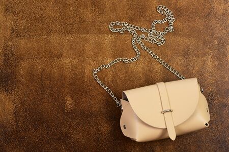 Female shoulder bag, close up with copy space. Beige handbag with long metal chain. Leather purse on brown suede background. Fashion concept.の写真素材