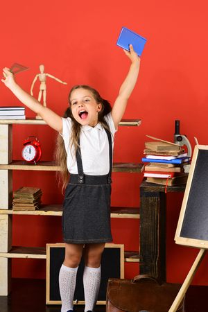 Back to school and education concept. Girl holds book and ruler near blackboard, copy space. Kid and school supplies on red background. Schoolgirl with happy face near bookshelf with school itemsの写真素材