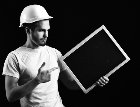 handsome bearded builder or foreman in white shirt and helmet on serious face holding board on black studio background, copy spaceの写真素材