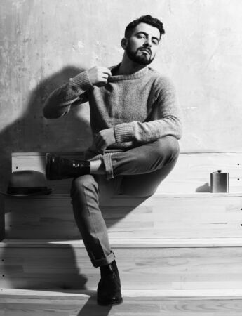 bearded man, long beard, brutal caucasian hipster with moustache in sweater sitting on wooden stairs with hat and flask with serious face on studio wall backgroundの写真素材