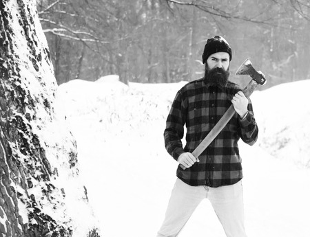 Handsome man or lumberjack, bearded hipster, with beard and moustache in red checkered shirt stands with axe in snowy forest on winter day outdoors on natural backgroundの写真素材