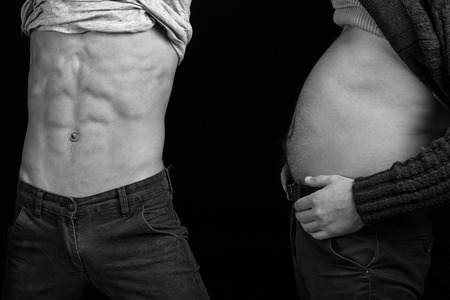 two friends showing paunchy and slim bellies with bare torso and six packs or abs in studio on black backgroundの写真素材