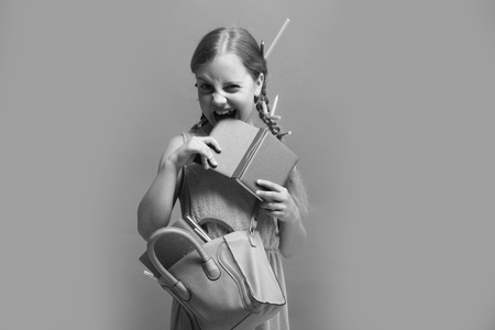 Girl with pink bag bites blue notebook. Kid with naughty face expression and colored pencils in hair. Pupil with long braids, isolated on blue background. Back to school and childhood conceptの写真素材