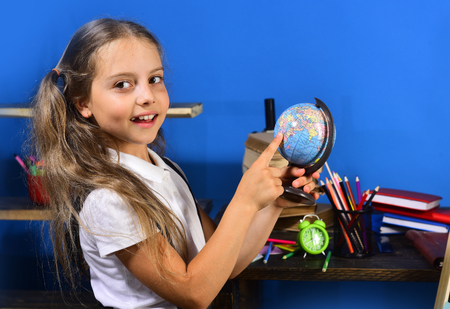 Girl stands by her desk with books. Kid and school supplies on blue wall background. Geography and back to school concept. Schoolgirl with curious and smiling face holds globe model in her classroomの写真素材