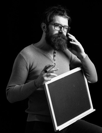 handsome bearded hipster man with long lush beard moustache and stylish hair on serious face in glasses yellow sweater holding board with cigar speaks by phone on black studio background, copy spaceの写真素材