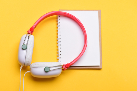 Music accessories and note taking concept. Modern and stylish earphones isolated on orange background, top view. Headset for music and blank page. Headphones in white and red color with empty notebookの写真素材