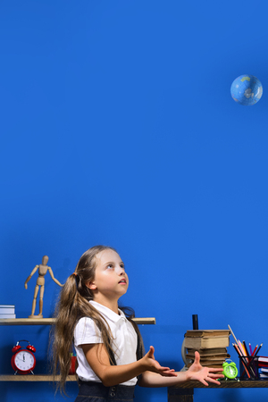 Girl in classroom with colorful stationery and books. Schoolgirl with scared face throws globe model up. Kid and school supplies on blue background, copy space. Back to school and geography conceptの写真素材