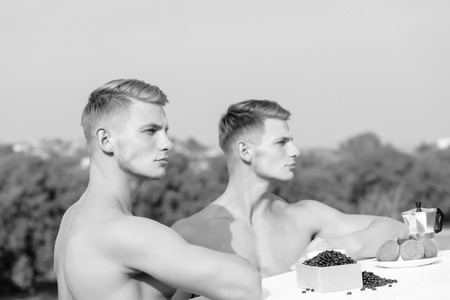 young handsome twins with sexy muscular athletic strong body has bare torso and strong belly having breakfast on roof with serious face on blue sky backgroundの写真素材