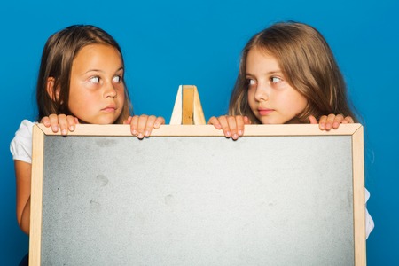 Children look at each other, copy space. Schoolgirls with serious faces stand behind blackboard. Girls in school uniform on blue background. Education and school concept.の写真素材