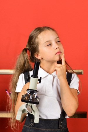 Girl holds microscope and thinks of experiment. Kid and school supplies on red wall background. Back to school and science concept. Schoolgirl with thoughtful face expression near bookshelfの写真素材