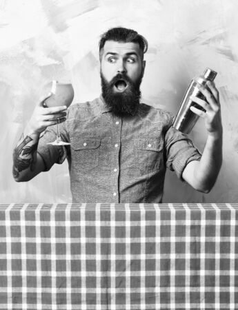 Bearded man, long beard. Brutal caucasian surprised hipster with moustache in denim shirt doing tropical alcohol fresh cocktail with shaker on blue checkered plaid on colorful texture backgroundの写真素材