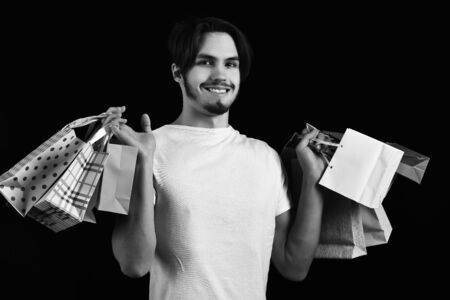 handsome bearded man in white shirt with stylish hair on serious face holding colorful package or bags on black studio backgroundの写真素材