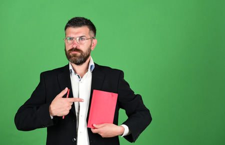 Knowledge and schedule concept. Professor with serious face expression. Notebook in red color in guys hands on green background. Teacher wears glasses and points at organizer. Man with beard and bookの写真素材