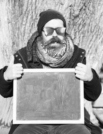 Handsome happy man, hipster, with beard and moustache in black sunglasses covered with white frost holds blank black board and thumbs up on winter day on natural background, copy spaceの写真素材