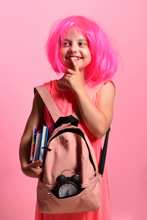 School girl with thoughtful face and smile wears backpack with black alarm on pink background. Girl holds books and markers. Back to school and education concept. Pupil in school uniform with pink wigの写真素材