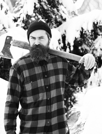 Handsome man or lumberjack, bearded hipster, with beard and moustache in red checkered shirt stands with axe in snowy forest on winter day outdoors on natural backgroundの写真素材