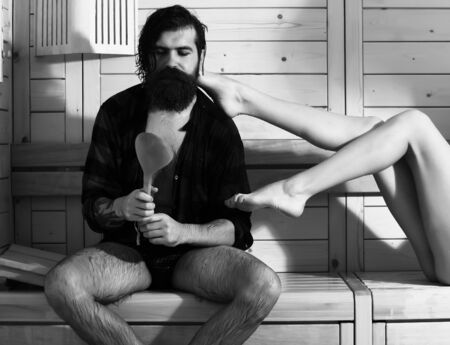 Handsome man or bather, bearded hipster with beard and moustache in red checkered shirt with spoon and sexy female legs wet of water in sauna, thermal bath on wooden backgroundの写真素材
