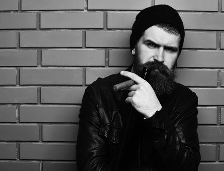 Bearded man, long beard. Brutal caucasian serious unshaven hipster holding bottle and smoking cigar with black leather jacket, hat and white shirt on brown brick wall studio background, copy spaceの写真素材