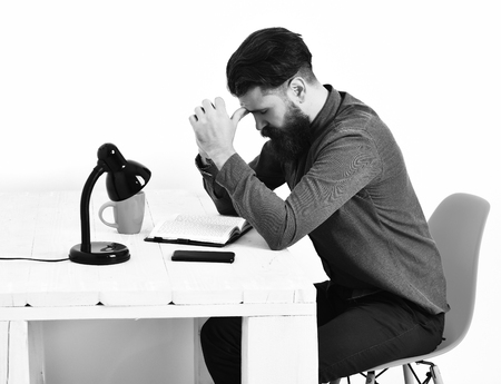 Bearded man, long beard. Brutal caucasian unshaven serious hipster with moustache sitting at white wooden table with black lamp, mobile phone and open notepad on it, isolated on white backgroundの写真素材