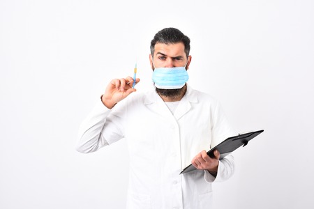 Medical doctor with stylish haircut holds black folder and syringe. Physician wears medical face mask isolated on white background. Doctor with beard dressed in white gown. Medicine and health conceptの写真素材