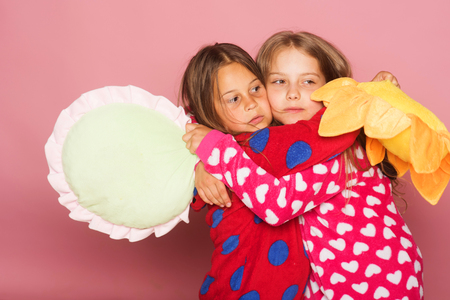 Childhood, friendship and happiness concept. Children with sleepy faces and loose hair hug. Girls in pink colorful polka dotted pajamas hold funny pillows. Kids stand on pink backgroundの写真素材
