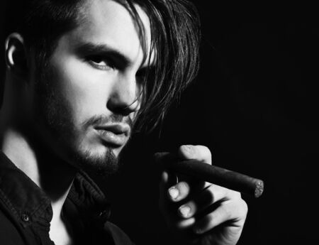 handsome bearded man in blue shirt with stylish hair on serious face holding cigar in black studio backgroundの写真素材