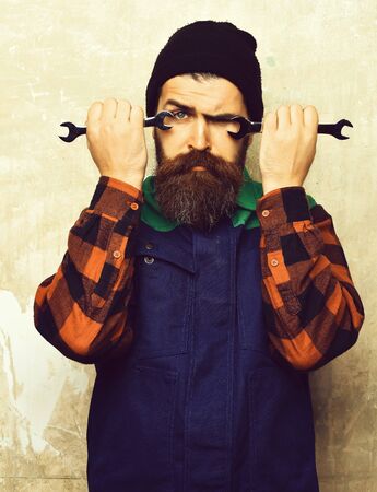 Bearded man, long beard. Brutal caucasian mechanic, unshaven hipster in black hat, checkered shirt and uniform holding metallic wrenches on texture studio backgroundの写真素材