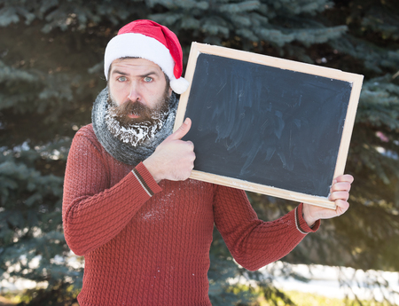 Handsome sad man in santa claus hat, bearded hipster with beard and moustache covered with white frost, holds blank black board on winter day outdoors on natural background, copy spaceの写真素材