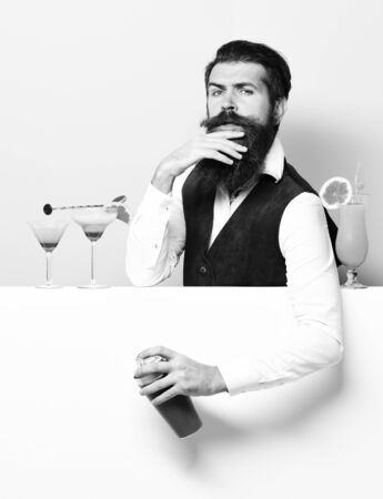 handsome bearded barman with long beard and mustache has stylish hair on serious face holding shaker and made alcoholic cocktail in vintage suede leather waistcoat on purple green studio backgroundの写真素材