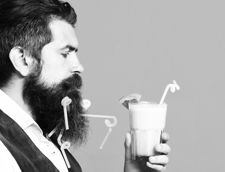 handsome bearded man with long beard and mustache has stylish hair on serious face holding glass of alcoholic beverage in vintage suede leather waistcoat on red studio backgroundの写真素材