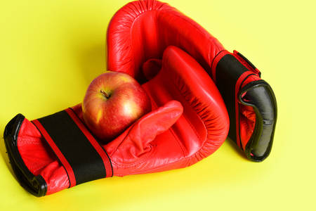 Boxing gloves in red color. Sport equipment and fruit isolated on bright yellow background. Pair of leather boxing sportswear with juicy red apple. Professional box fight and dieting conceptの写真素材