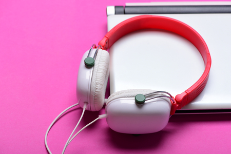 Earphones in red and white colors made of plastic with computer. Music and digital equipment concept. Electronics isolated on pink background. Headphones and silver laptop. Sound recording ideaの写真素材