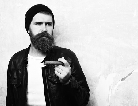 Bearded man, long beard. Brutal caucasian serious unshaven hipster smoking cigar with black leather jacket, hat and white shirt on battered wall background, copy spaceの写真素材