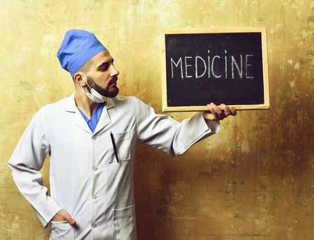 Medicine word written on blackboard which is held by bearded doctor with serious face expression on beige backgroundの写真素材