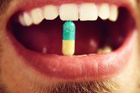 Patients teeth hold transparent capsule, close up. Guys mouth with long blue and yellow drug, defocused. Medical treatment or healthy choice conceptの写真素材