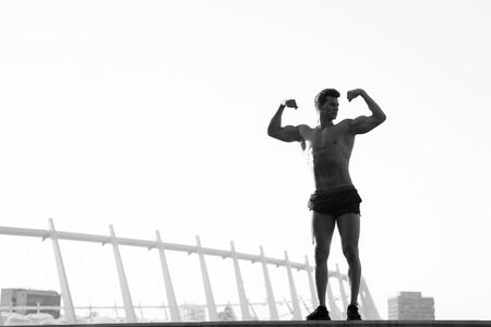Man with muscular body on clear sky. Sportsman flexing arms on roof. Fitness and sport. Athlete training outdoors. Healthy lifestyle concept, black and whiteの写真素材