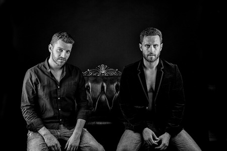 two young handsome bearded muscular men sexy macho with beard on serious face in stylish jacket and shirt posing in studio sitting on leather chair, black and whiteの写真素材