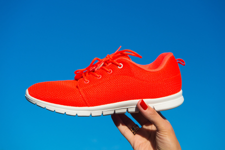 Sports shoes sneakers on blue sky background. Sports in the open air. woman hand holding orange sneakers fitness and healthの写真素材