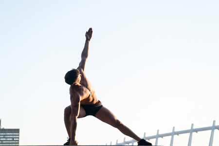 Man stretching hands and legs on roof. Sportsman with muscular body on white sky. Athlete doing workout outdoors. Fitness and sport. Healthy lifestyle concept, copy spaceの写真素材