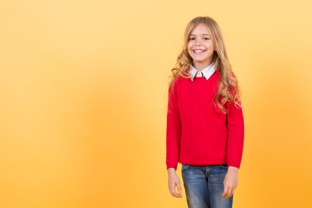 Child girl with long curly blond hair happy smile in red sweater and blue jeans on orange background. Fashion, style, beauty, look concept, copy spaceの写真素材