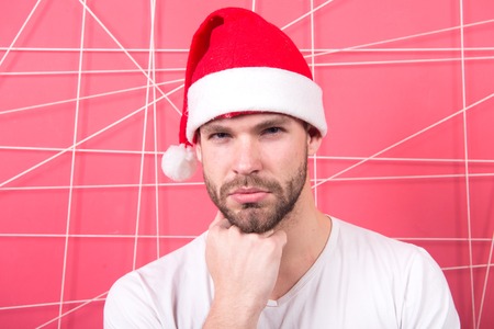 Macho in santa hat touch beard on serious face on pink background. Barbershop, grooming, male beauty. Christmas, new year, winter, holidays, celebration, party conceptの写真素材
