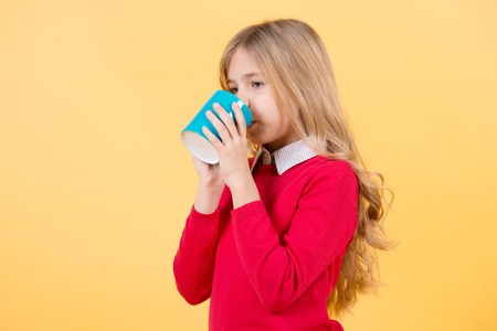 Girl drink tea or coffee from cup with long blond hair in red sweater on orange background. Thirst, dehydration concept.の写真素材