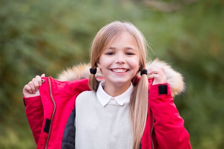 Girl with blond hair ponytail smile on natural landscape. Child in red coat on idyllic autumn day. Kid fashion and style. Happy childhood concept. Beauty, look, hairstyle.の写真素材