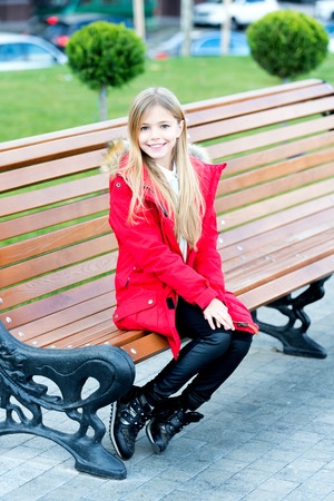 Girl in red coat sit on bench in park. Kid enjoy autumn day. Happy childhood concept. Child with blond long hair smile outdoor. Leisure, relaxation, lifestyle.の写真素材
