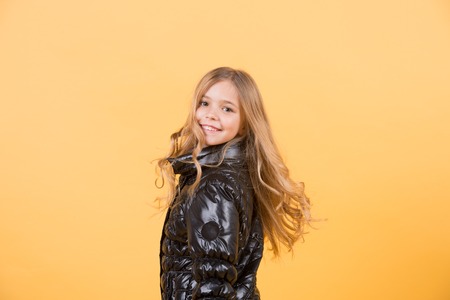 Fashion, autumn style, trend. Girl in black coat on orange background. Happy childhood concept. Child model smile with long blond hair. Kid beauty, look, hairstyle, copy spaceの写真素材